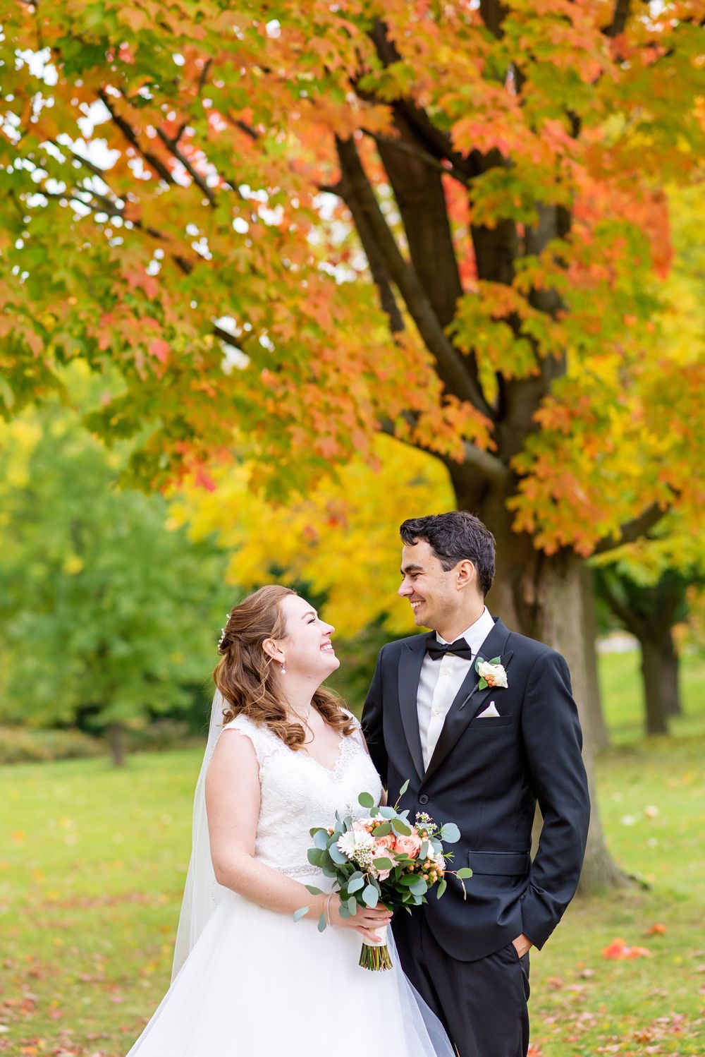 Newlyweds share a romantic moment under vibrant autumn trees with orange and green foliage.