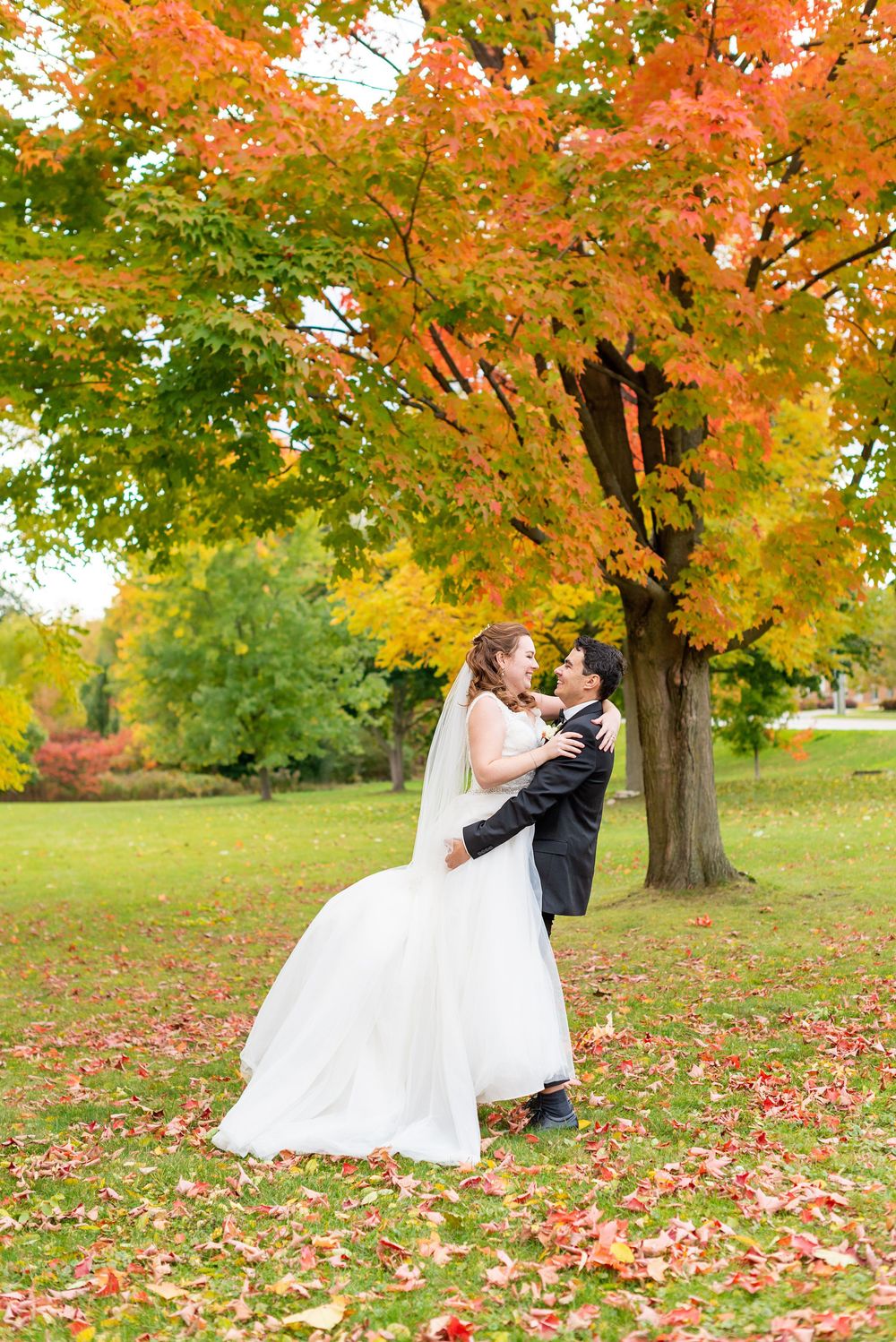 A couple shares a romantic moment under vibrant autumn maple trees with fallen orange leaves scattered on the ground.