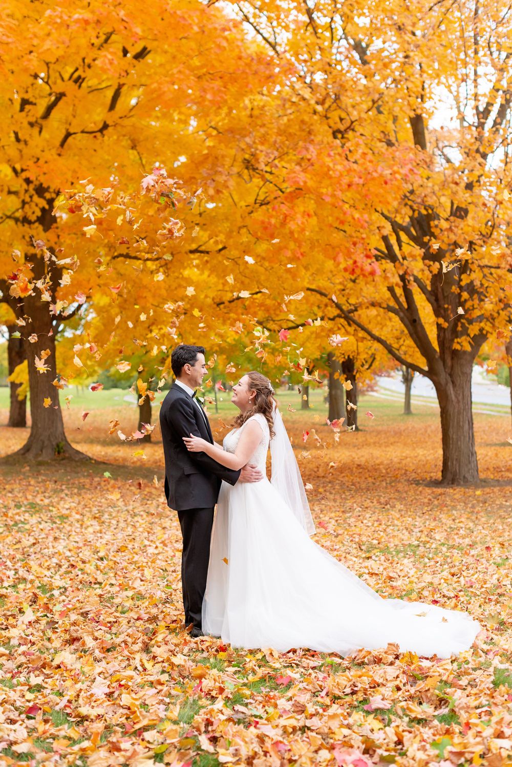 A couple shares a romantic moment surrounded by vibrant golden autumn trees and fallen leaves in a park setting.