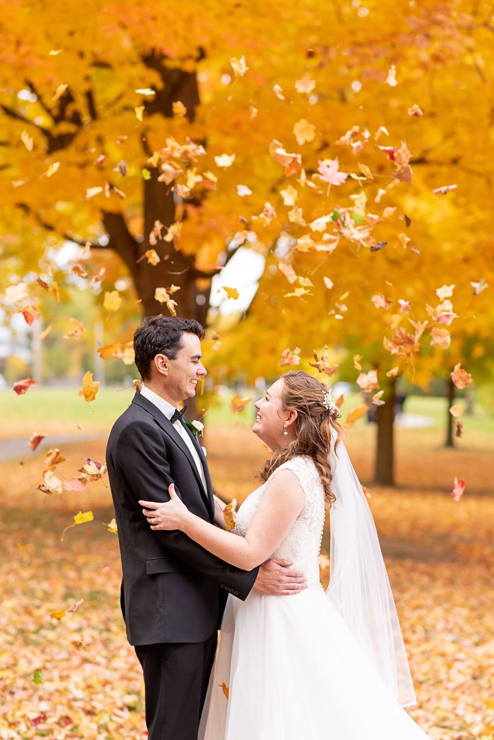Couple in wedding attire dances among falling autumn leaves in a park with golden foliage.
