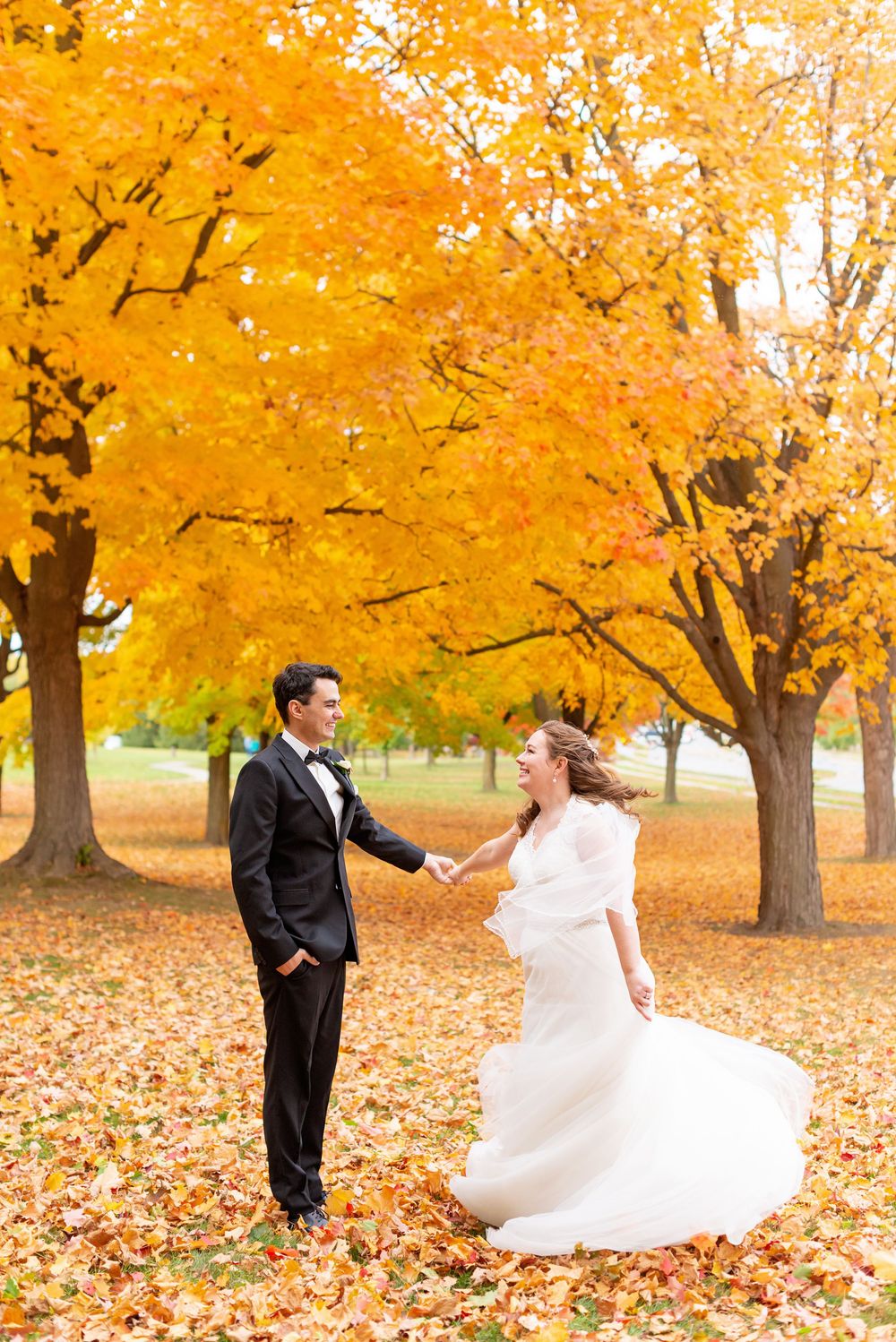 A couple dances playfully among vibrant yellow autumn trees with fallen leaves scattered on the ground.
