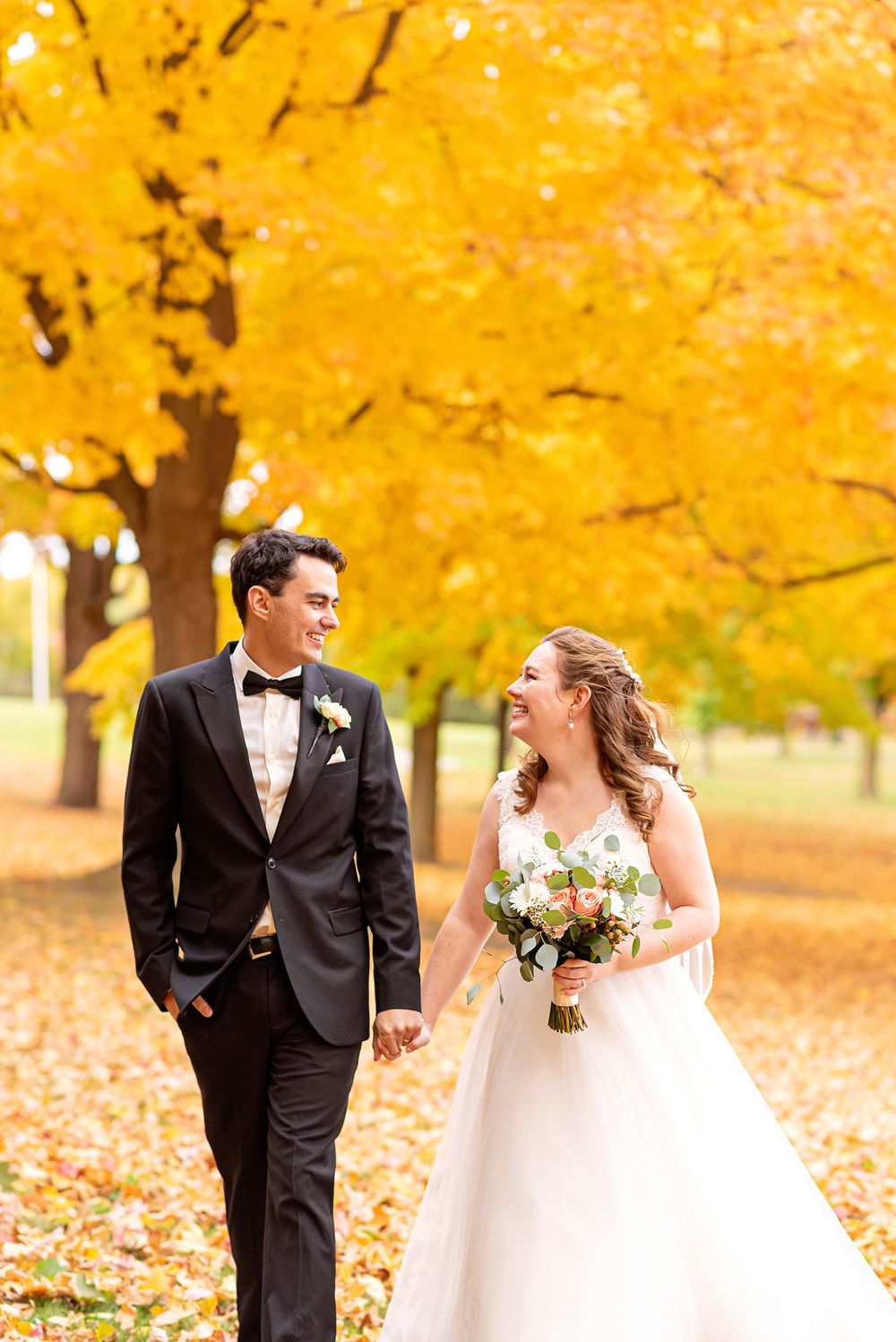 A couple in wedding attire walks together on an autumn day with vibrant yellow fall foliage in the background.