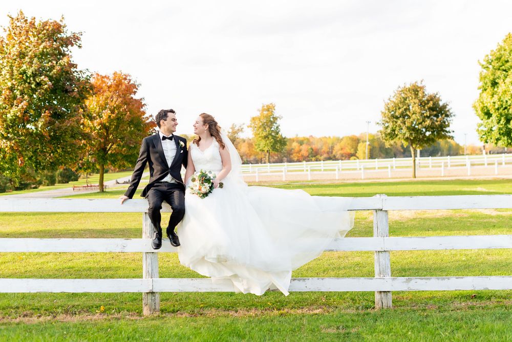 A couple in wedding attire sit playfully on a white fence surrounded by fall foliage.