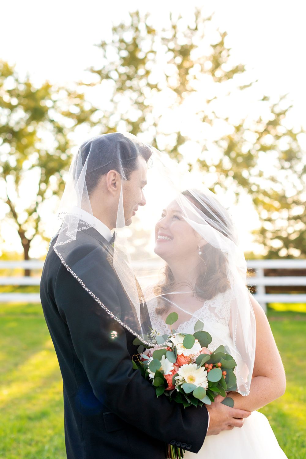 A romantic wedding portrait backlit by golden sunlight with white fencing and greenery in the background.