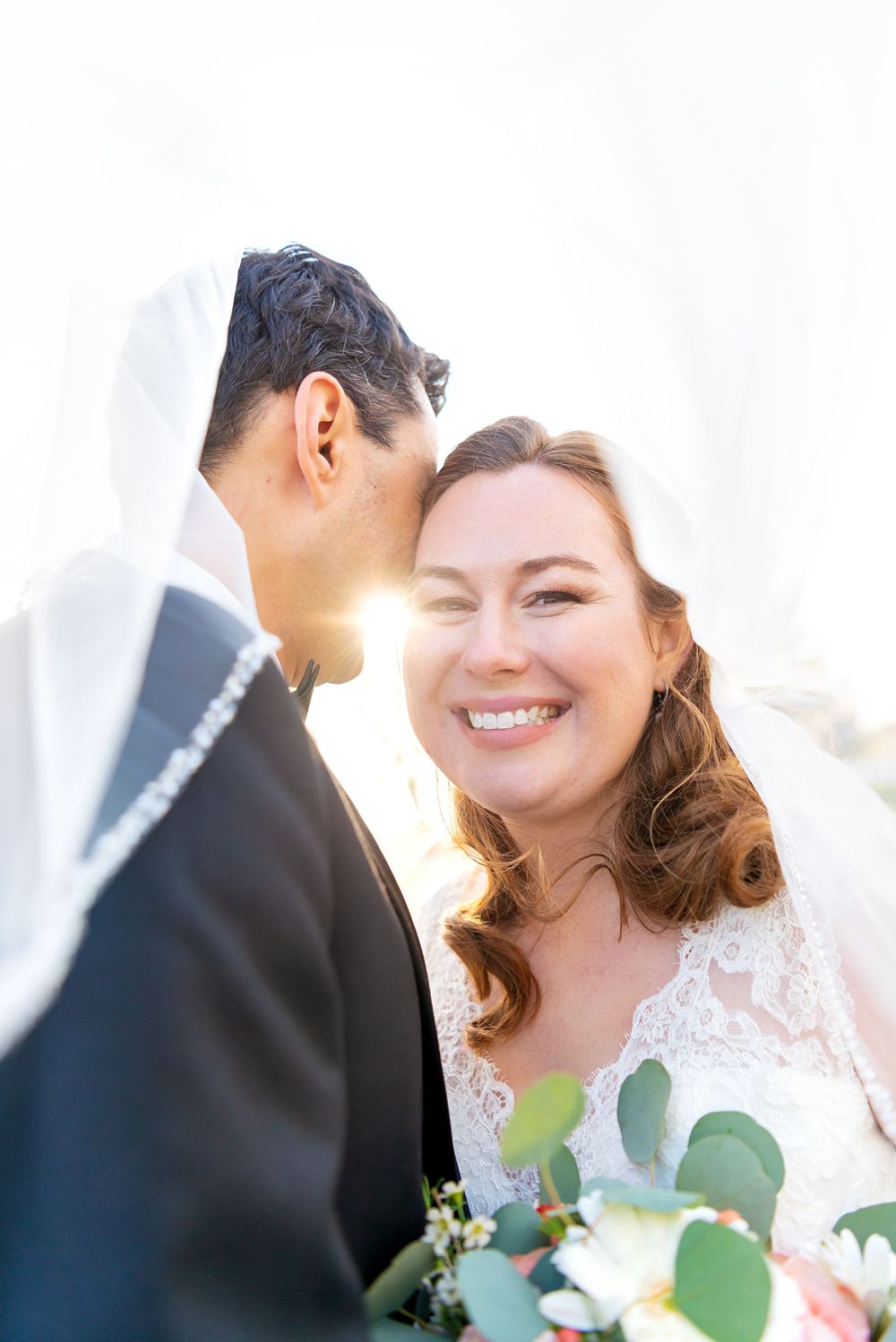 A happy couple shares a tender moment together under a bridal veil with greenery in the background.