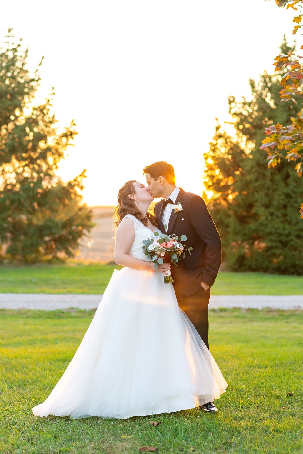 A bride and groom share a romantic kiss at sunset on a grassy lawn with trees silhouetted in the golden light.