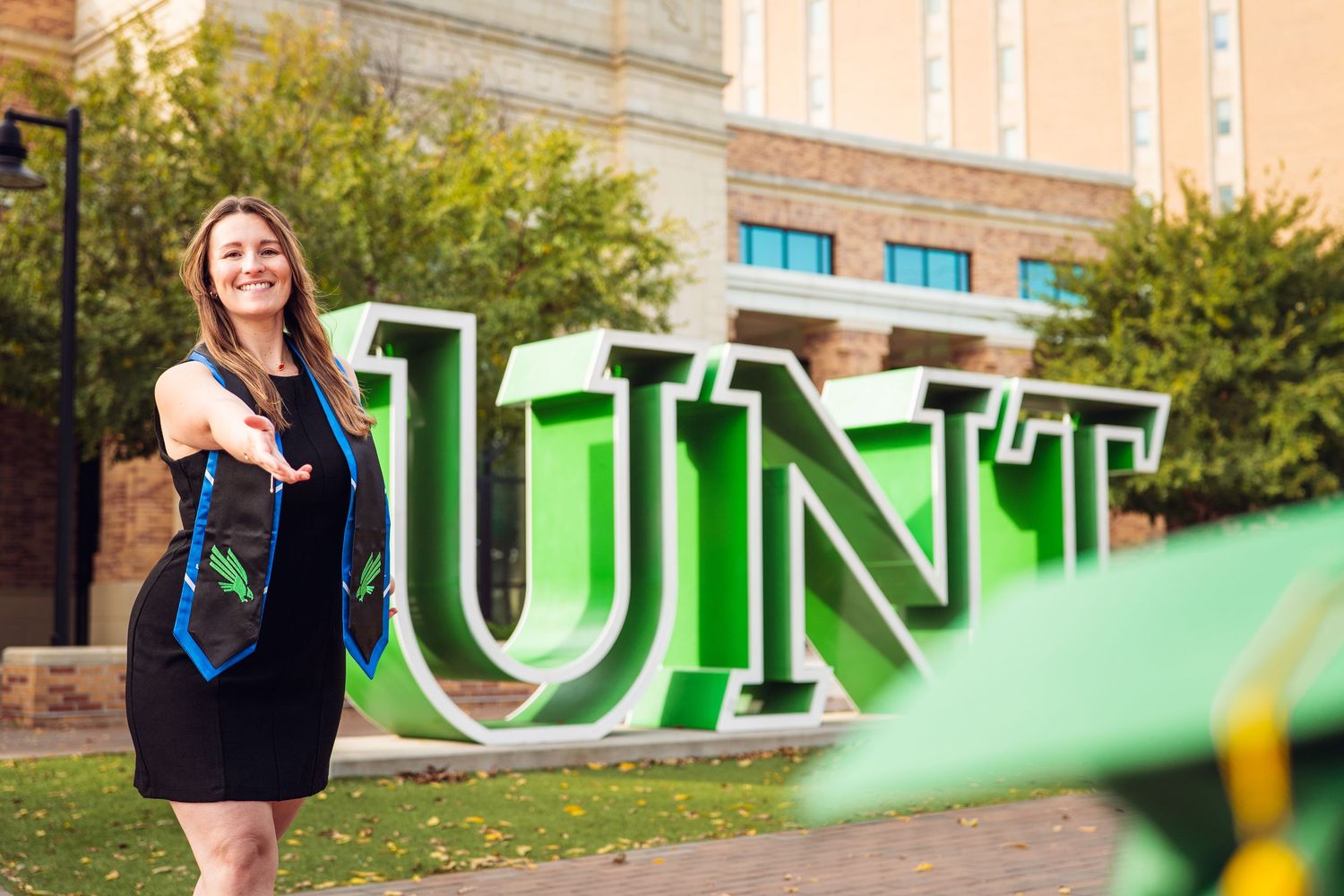 A person poses in front of large green UNT letters on a university campus.