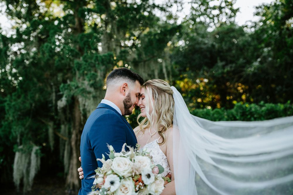 A wedding couple embraces outdoors with a flowing veil in a romantic garden setting.