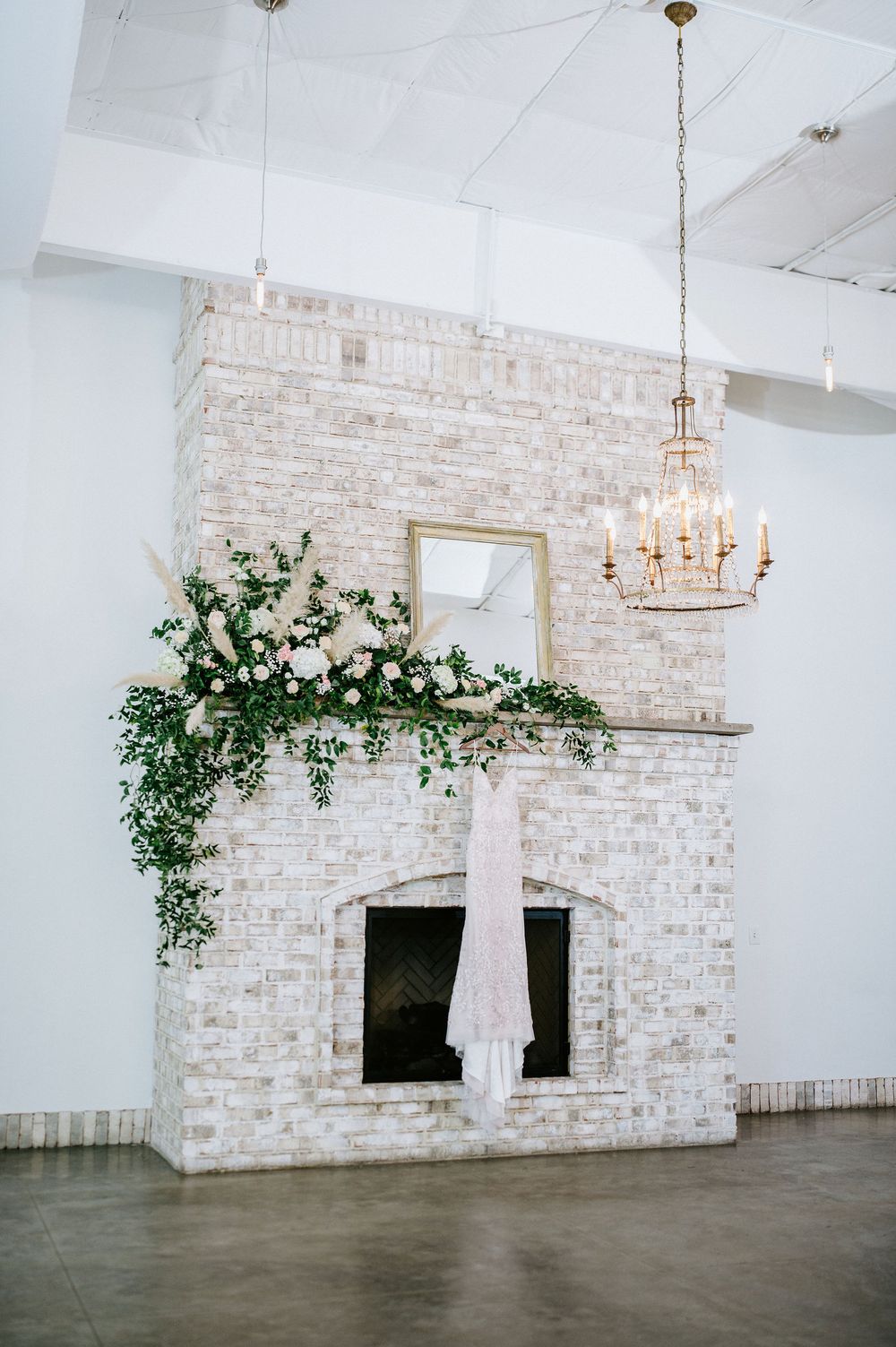 White wedding dress hangs elegantly in modern venue with whitewashed brick walls, chandelier, and cascading greenery decor.