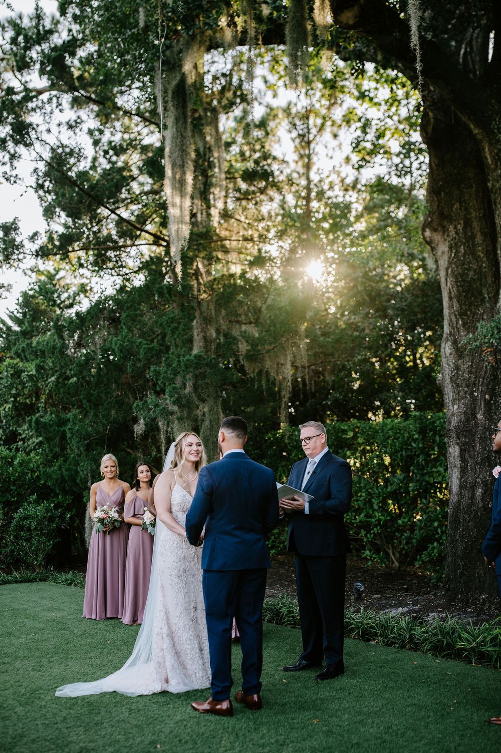 An outdoor wedding ceremony takes place at sunset under moss-draped oak trees in a lush garden setting.