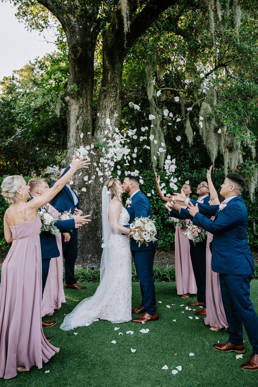 Wedding party celebrates outdoors under moss-draped oak trees as flower petals float through the air.