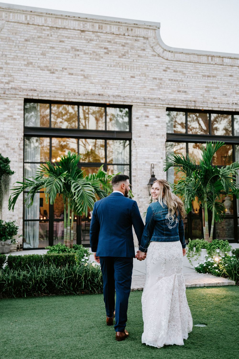 A couple walks together on a manicured lawn outside a modern brick building with large windows and tropical landscaping.