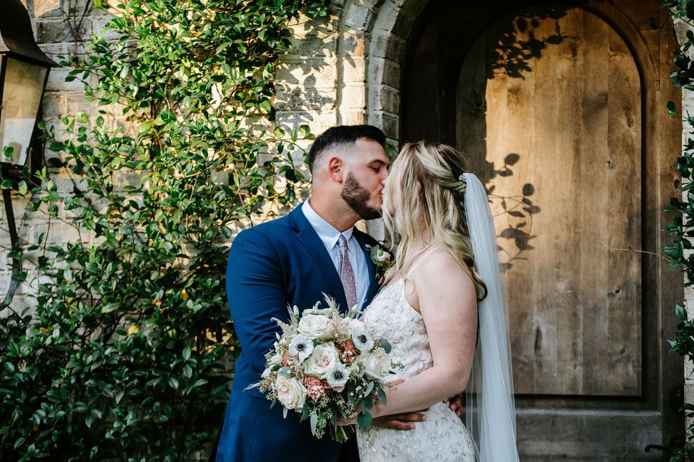 A romantic wedding moment under an outdoor arch decorated with white floral arrangements and greenery.