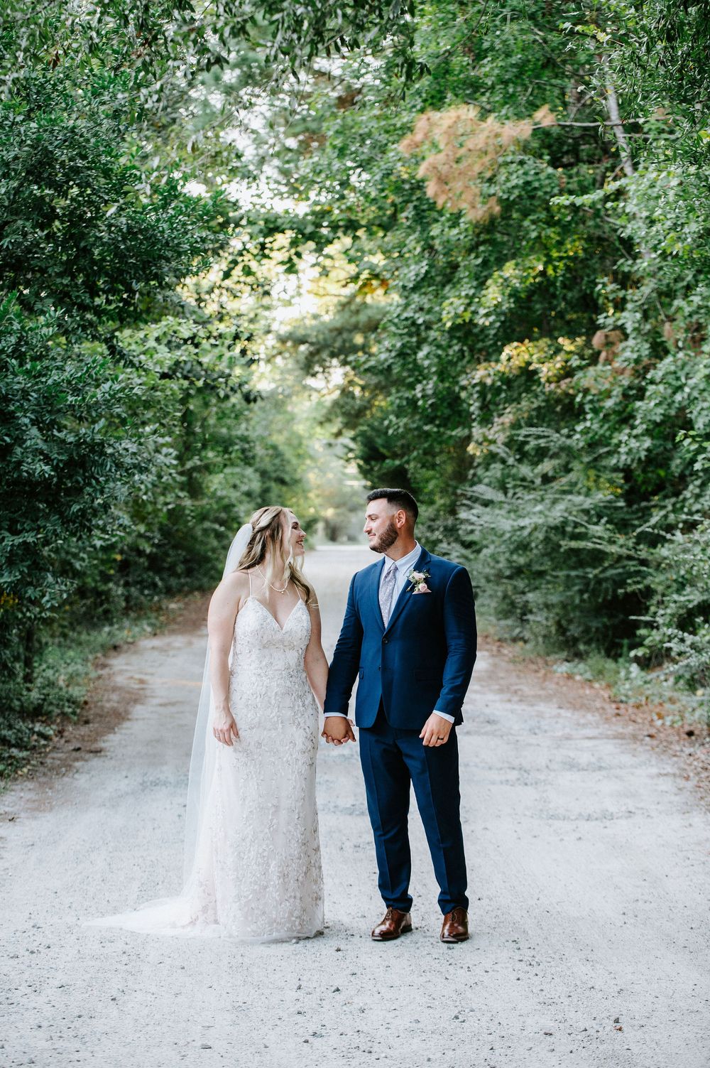 A couple walks hand in hand down a tree-lined gravel path during their wedding day.