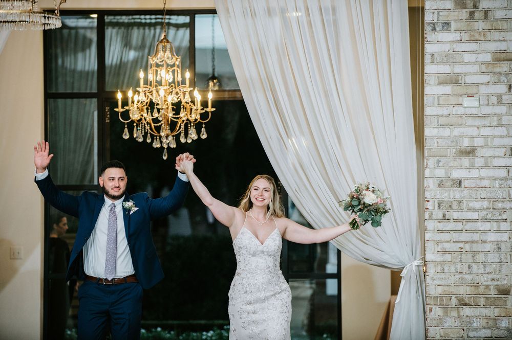 A couple makes their grand entrance into an elegant reception hall with crystal chandeliers and white drapes.