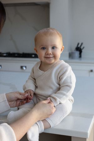 Smiling baby in soft tones sits on a counter smiling at the camera during a family photography session in Brighton