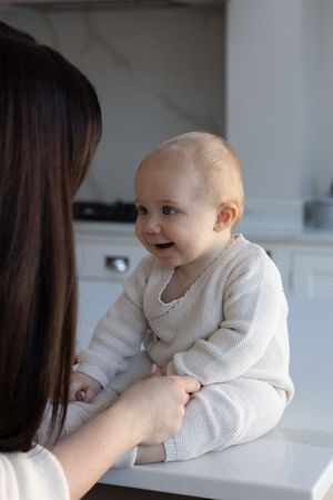 A smiling baby in a cream knit outfit sits on a counter facing her mother during a family photography session in Brighton