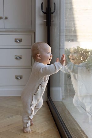 Curious baby standing barefoot, touching glass door reflection in bright modern home in Brighton, taken during a family photography session