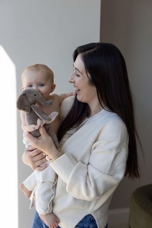 Smiling mother holds her baby and a stuffed elephant toy in a bright naturally lit room, photographed during an in home session in Brighton