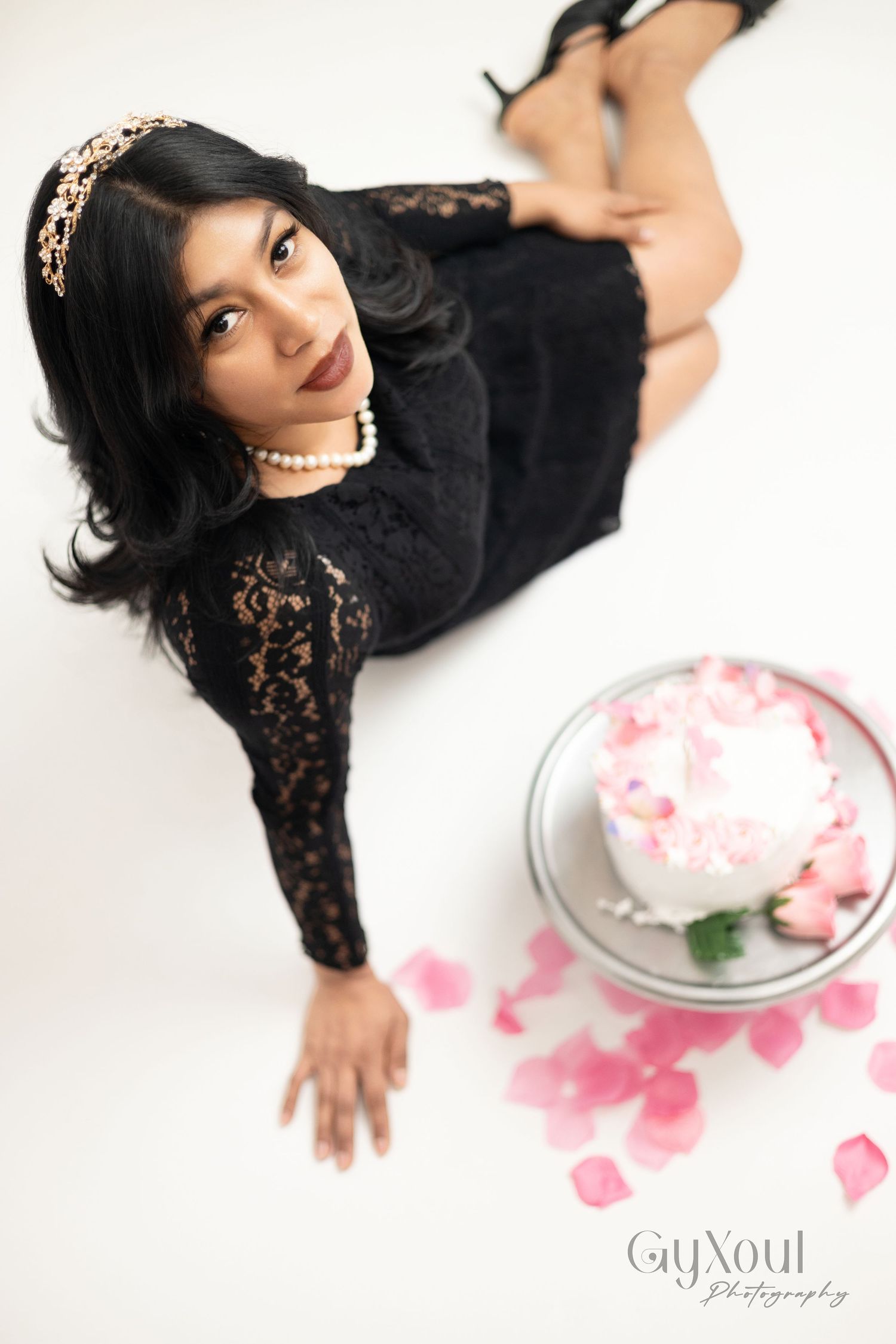 Elegant woman in black lace dress poses with pink rose petals scattered on white surface in artistic photo series.
