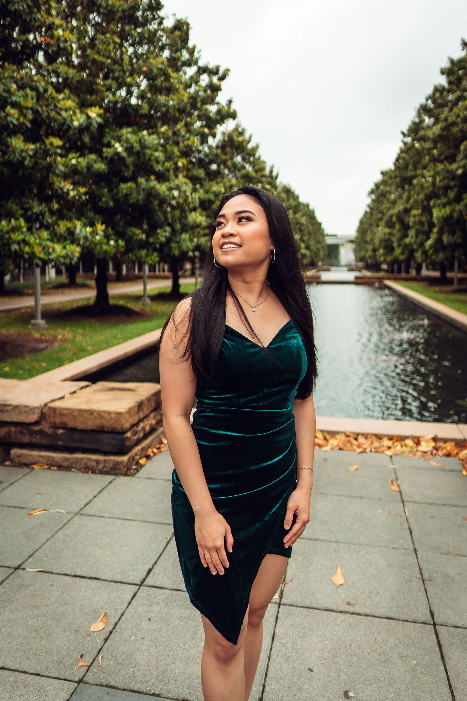 Student in emerald velvet dress poses elegantly by campus waterway. UTD Graduate Portraits by Eric Aaberg Photography in DFW.