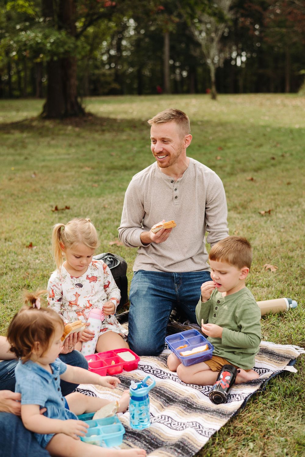 A family enjoys a picnic together on a striped blanket in a grassy park setting.