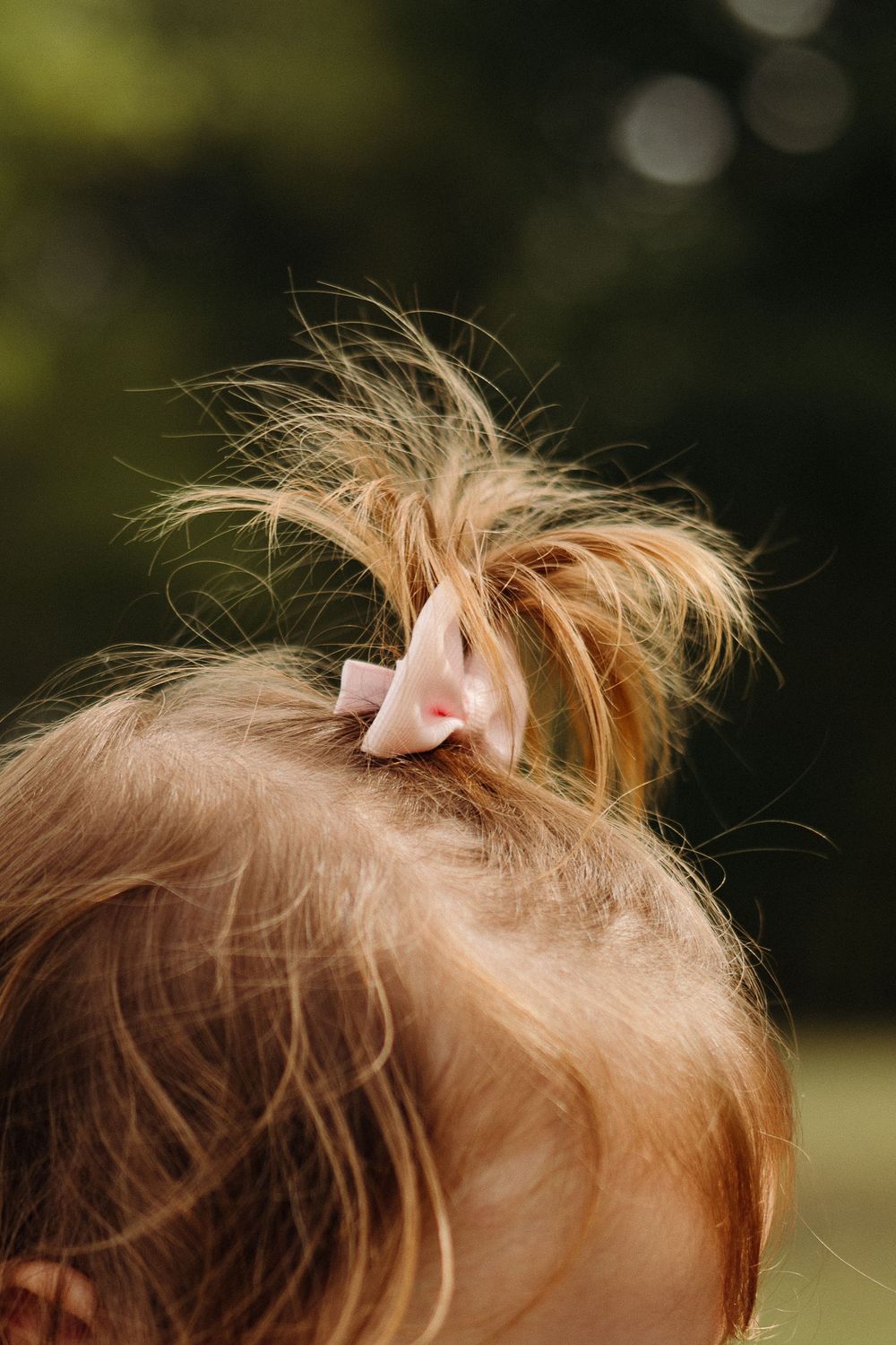 Close up view of a small messy blonde hair bun with a pink bow against a blurred green outdoor background.