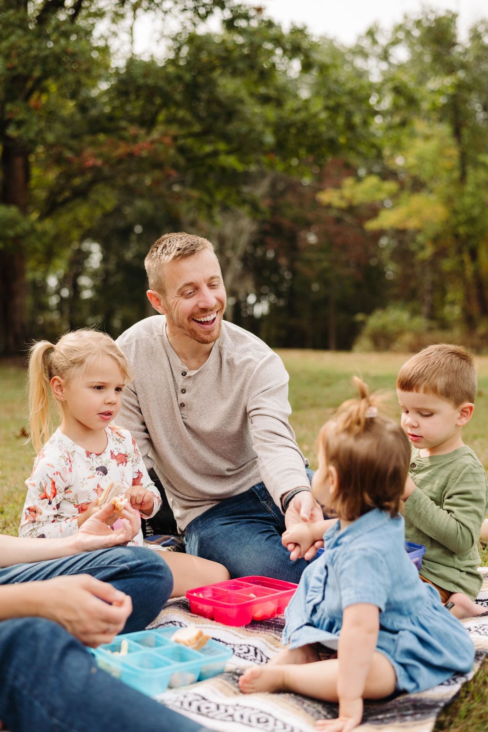 Family enjoys playful moments together on a striped blanket in a sunny park setting.