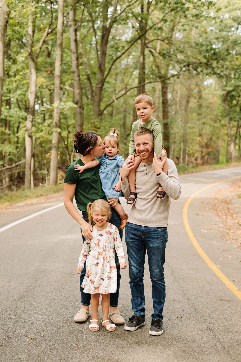 A family stands together on a curved forest road during autumn with tall trees in the background.