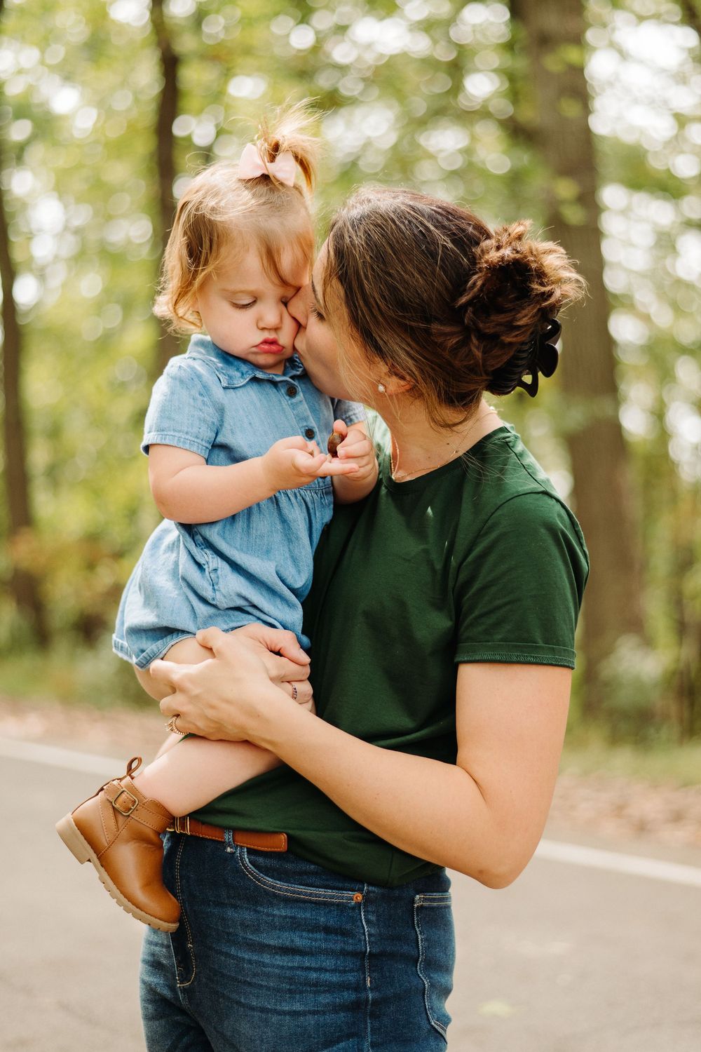 Parent and toddler share tender moments together in a wooded outdoor setting wearing casual denim and green attire.