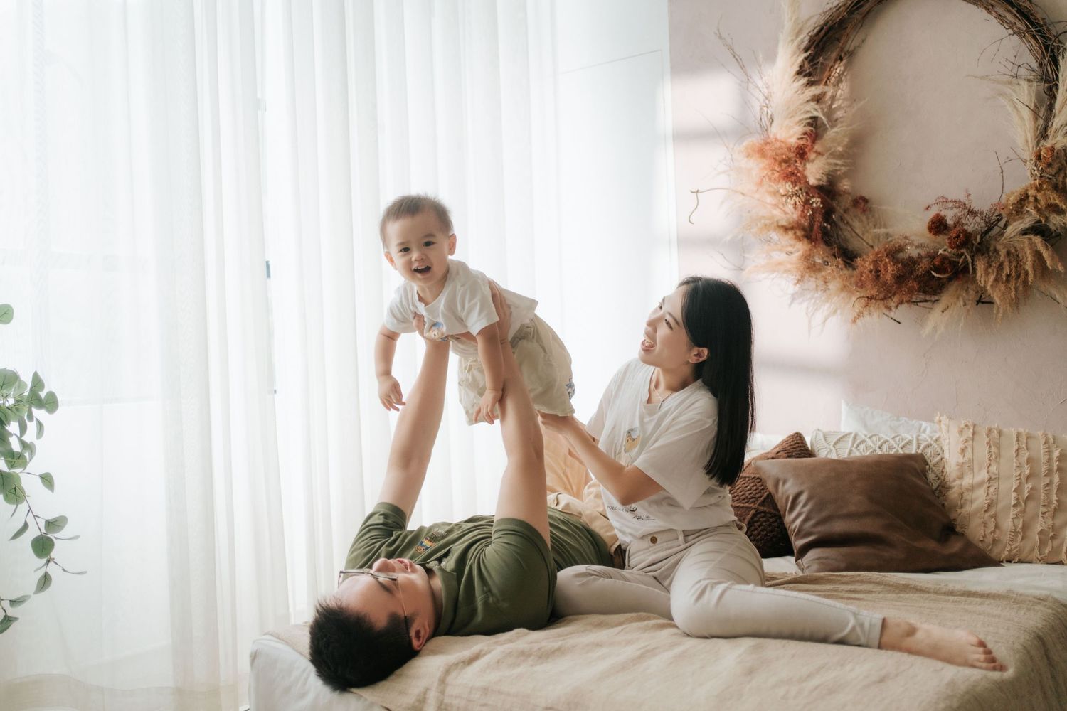 Happy family playing with baby on bed, parents lifting laughing infant in bright cozy bedroom with boho decor.幸福的一家人在床上和寶寶玩耍，父母抱著咯咯笑著的嬰兒