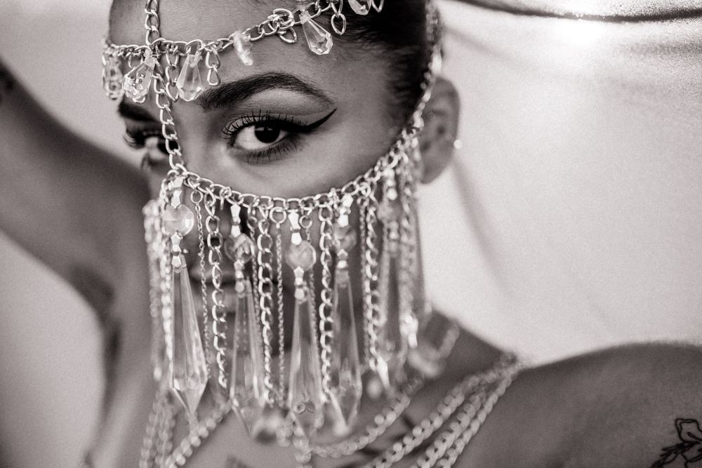 Black and white close-up of a woman in an ornate beaded face veil with crystal pendants, bold winged eyeliner, and layered chain jewelry.