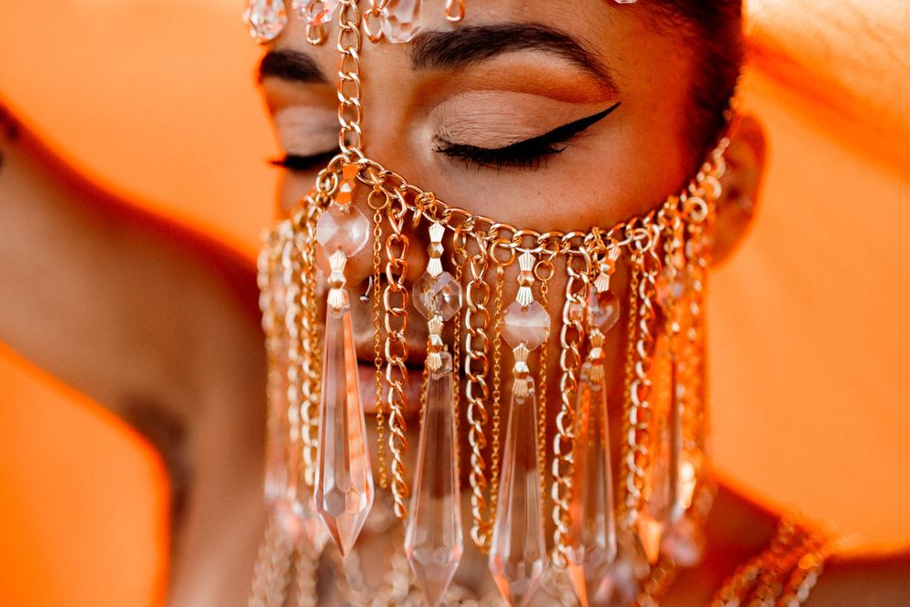 Close-up of a woman wearing an ornate beaded face veil with dangling crystal drops, dramatic winged eyeliner, and a warm orange background.