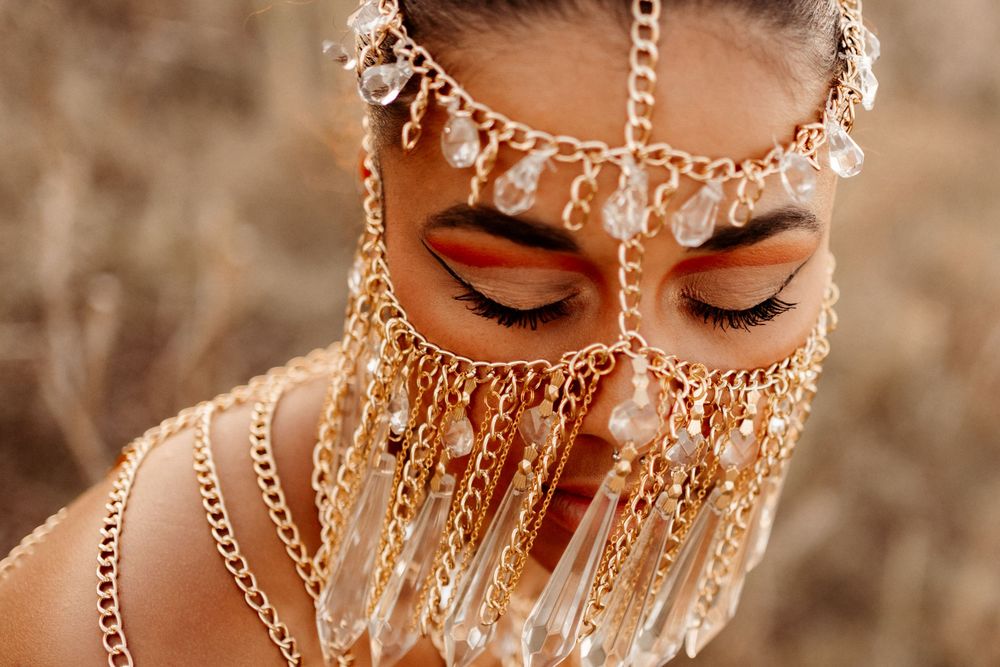 Woman with closed eyes wearing an ornate gold face chain veil with crystal drops and orange eyeshadow against a golden field background.