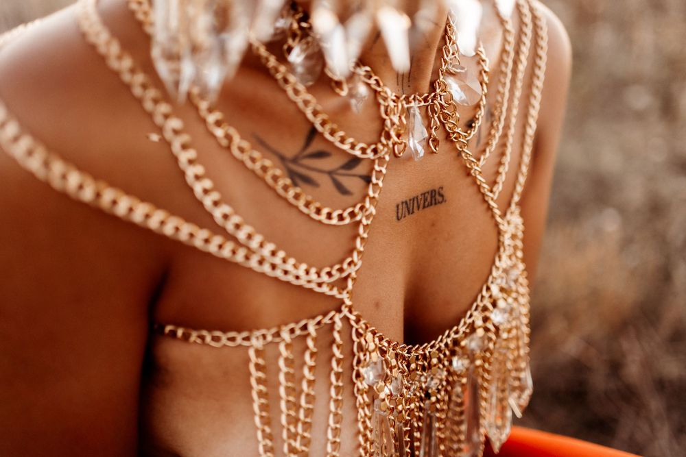 Close-up of a woman wearing layered gold chain body jewelry with crystal pendants and a small chest tattoo, in warm golden outdoor light.