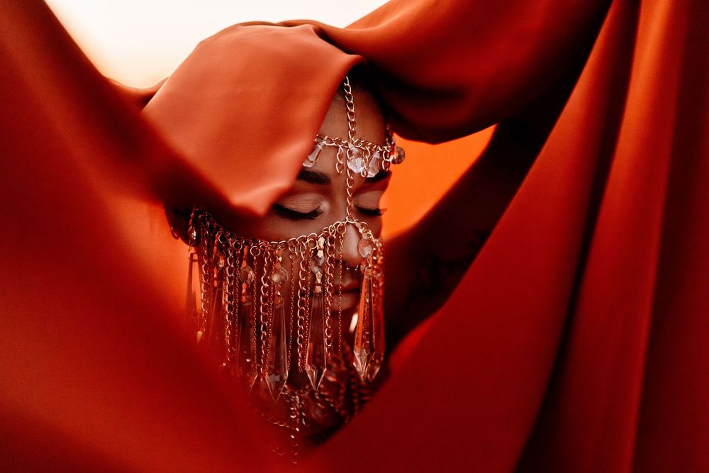 Ornate beaded crystal tassel hangs elegantly against flowing red fabric drapes in dramatic lighting.