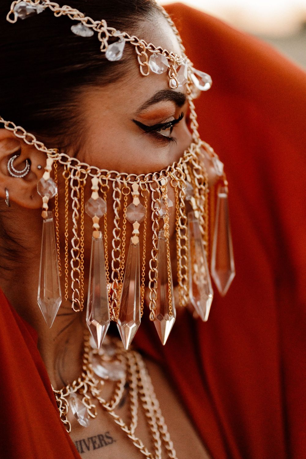 Intricate gold chain headpiece with crystal pendants against a red background showcasing dramatic bronze eye makeup.