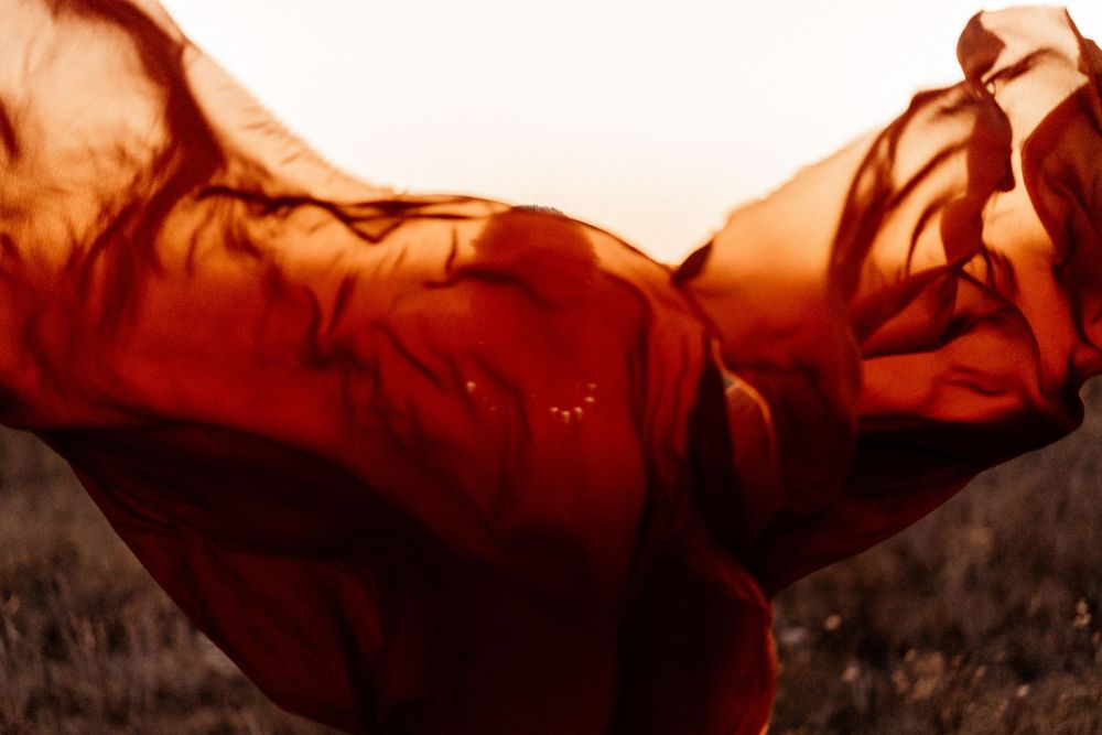 Flowing red-orange fabric billows in the wind against a soft golden sky and blurred grassy field at sunset.