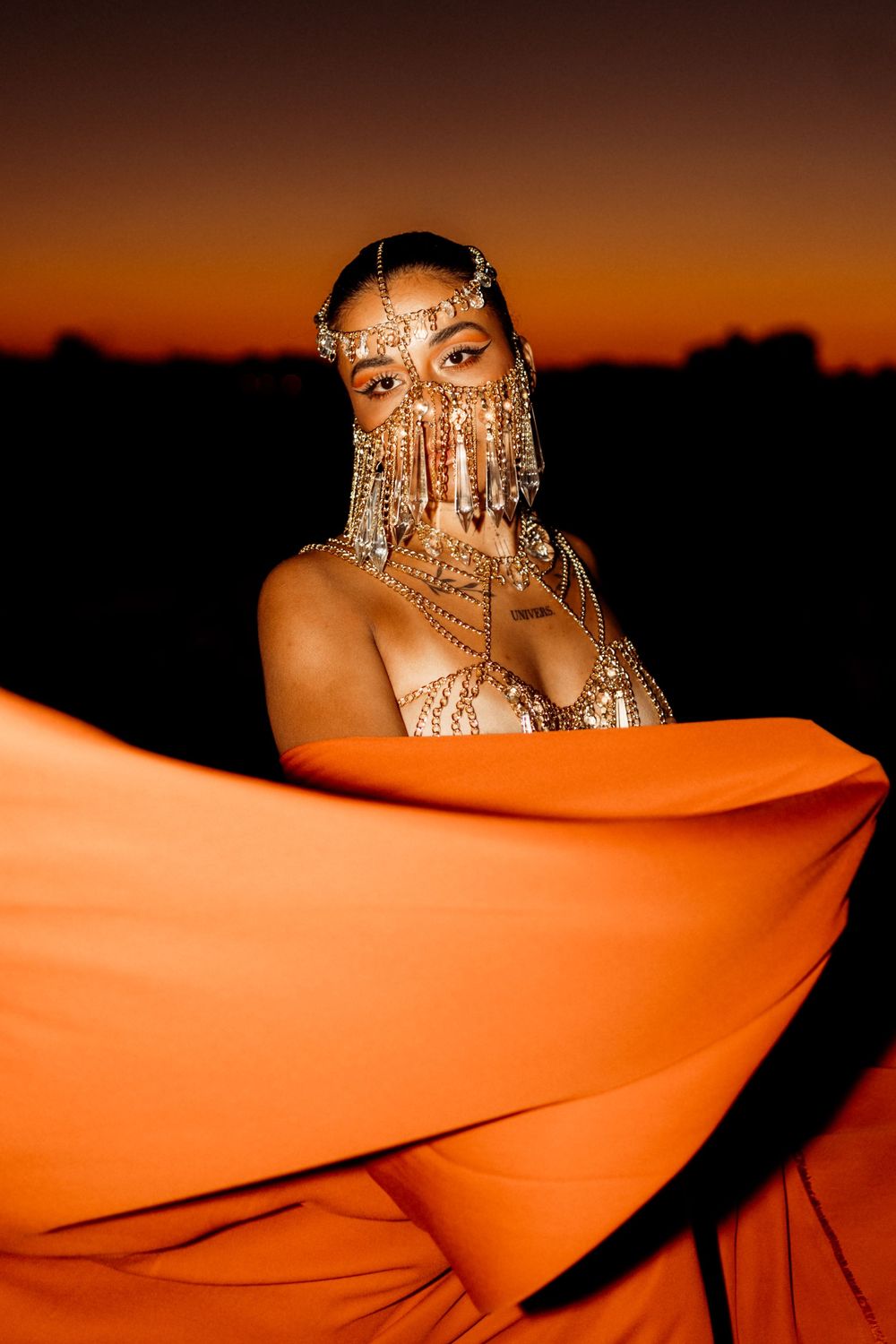 A person in elaborate gold jewelry and face decorations poses against a dramatic orange sunset sky backdrop with flowing fabric.