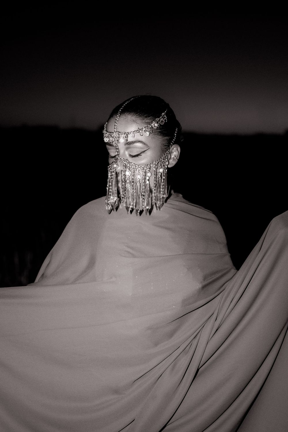 A dramatic black and white portrait showing an ornate beaded face veil and headpiece against a dark background.