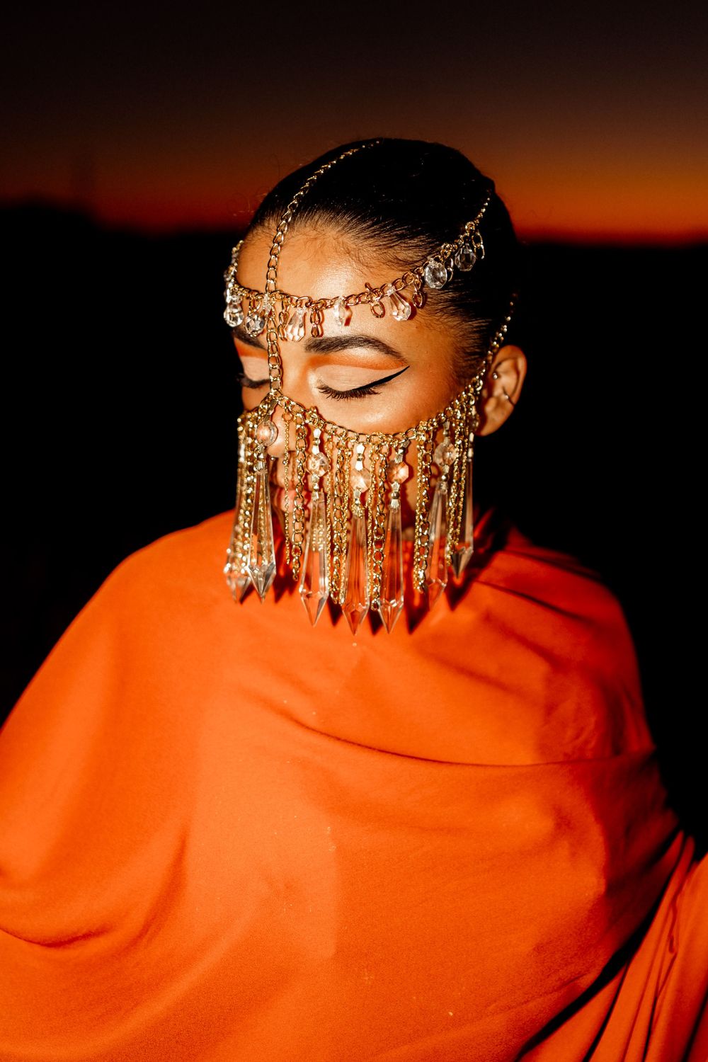 Figure in vibrant orange fabric wearing an ornate golden headpiece with cascading jewels against a sunset sky.