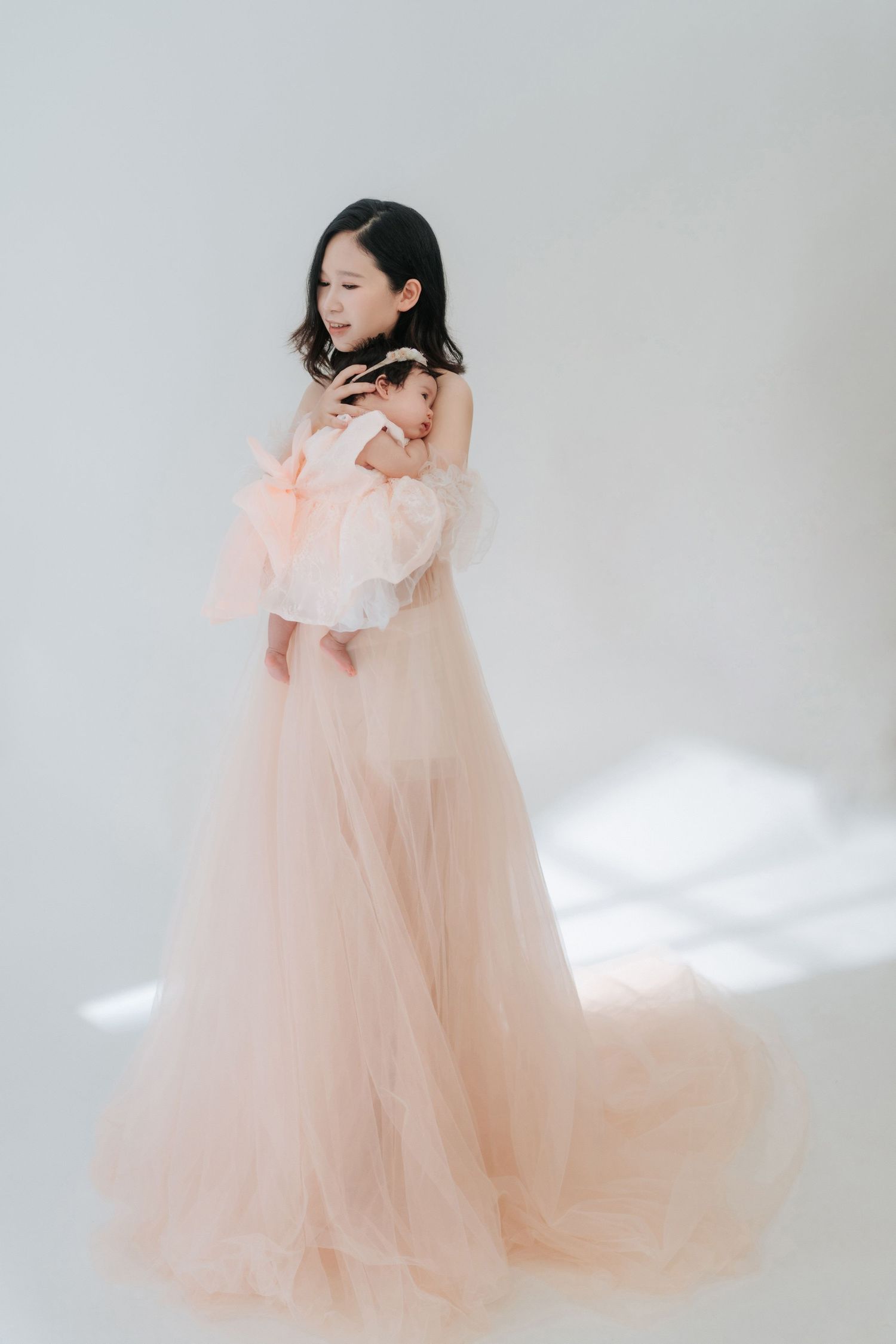 A figure in a flowing peach-colored tulle gown stands elegantly against a white backdrop in a dreamy studio setting.在夢幻般的攝影棚環境中，一位身著飄逸的蜜桃色薄紗