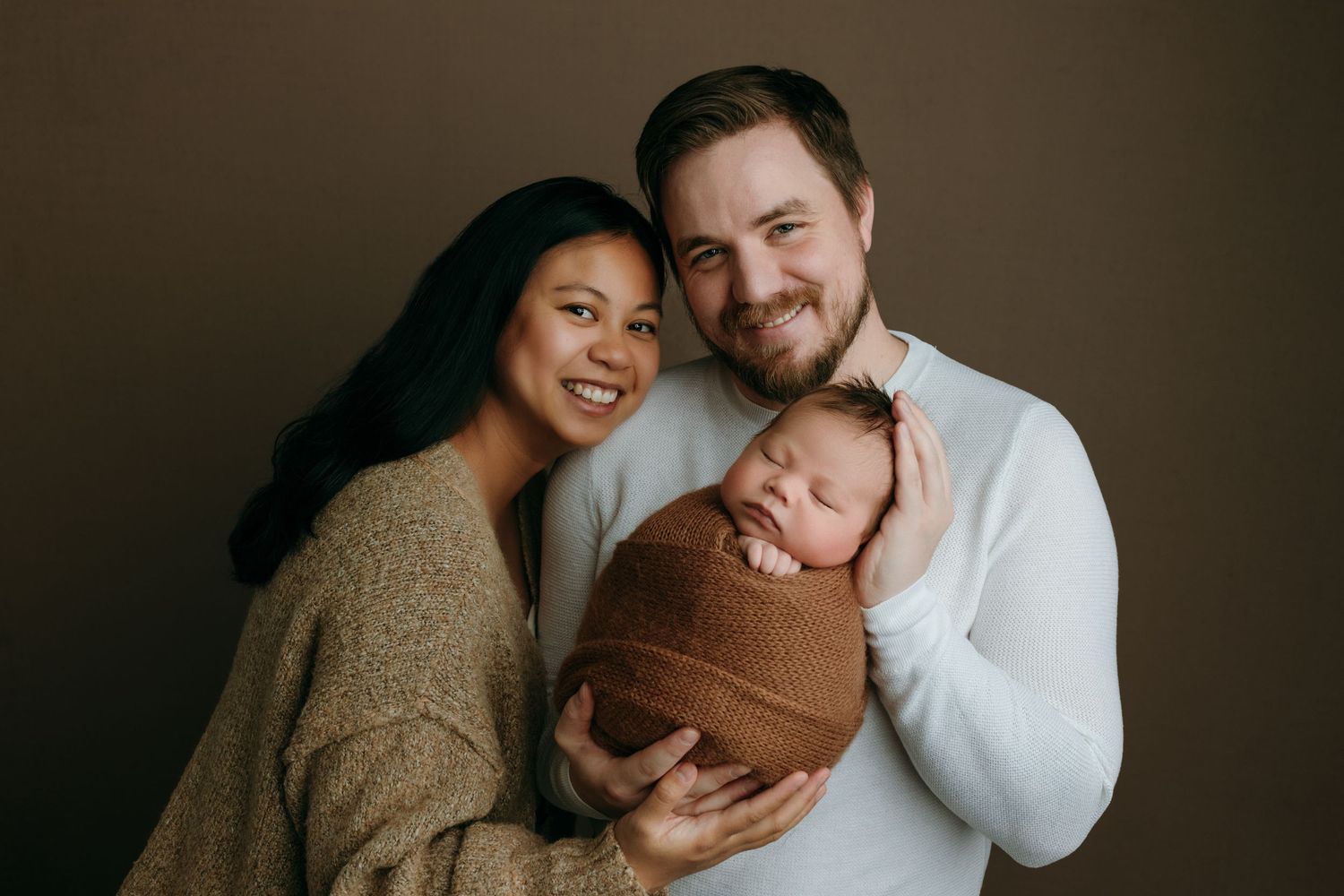A newborn family poses together for a portrait against a dark background, with a baby in brown at the family photo studio in Victoria BC