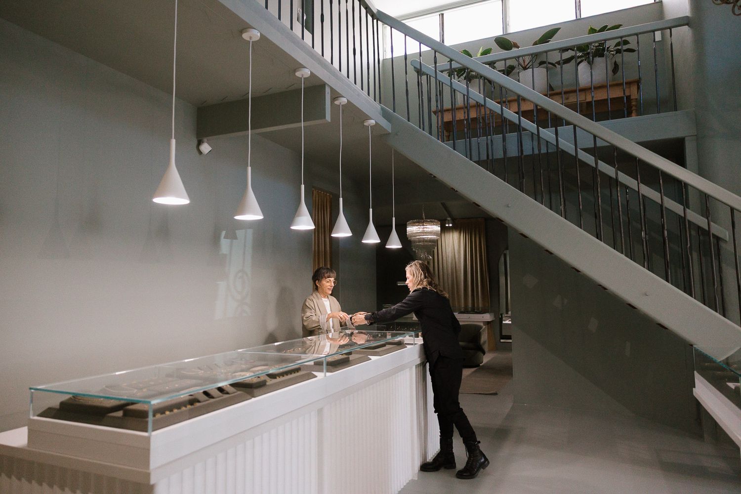 Two people at a modern jewelry display counter near a staircase in a contemporary retail store.