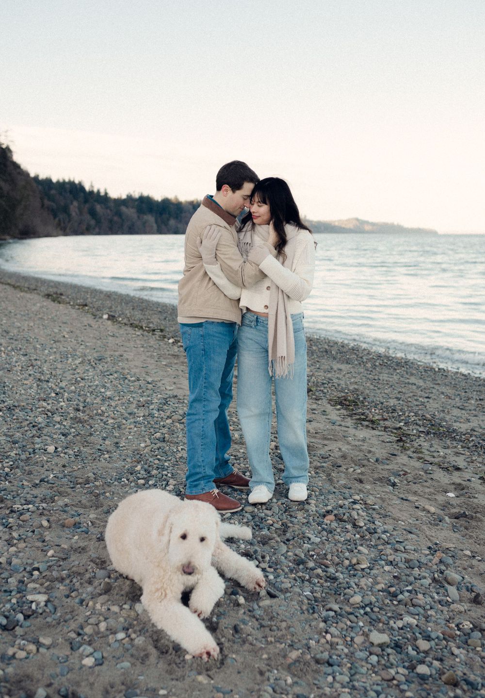 A couple and their white dog share a romantic moment on a rocky beach at sunset.