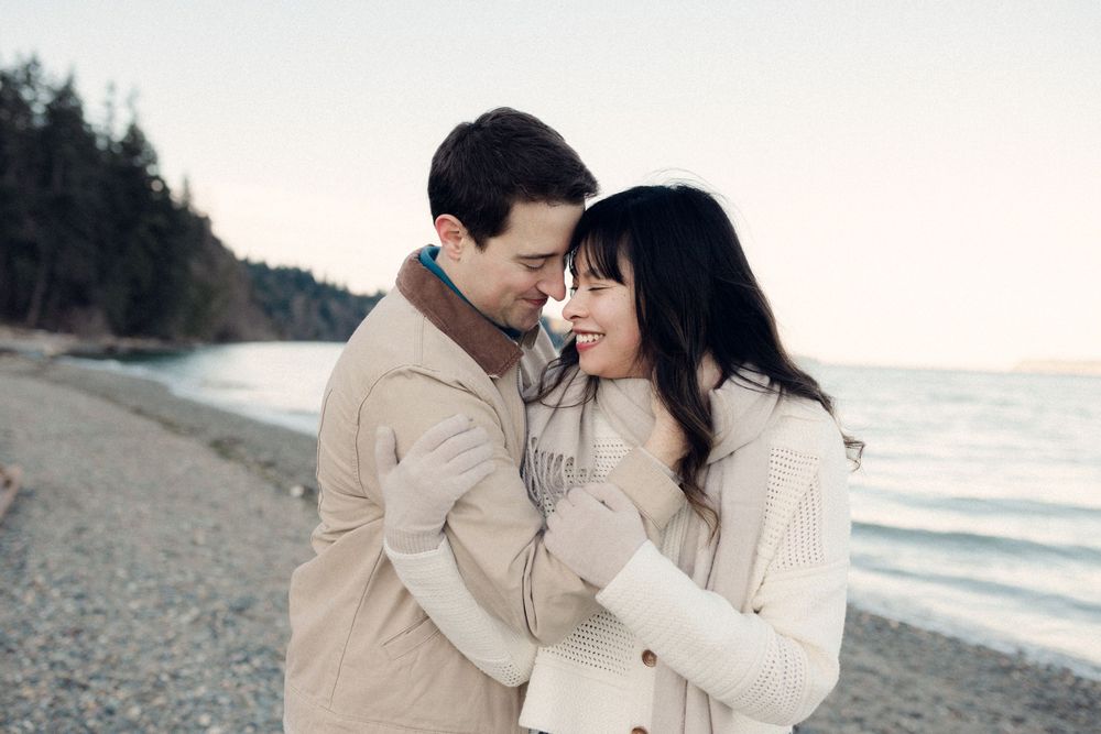 Romantic couple in cream sweaters embracing on misty beach at sunset.