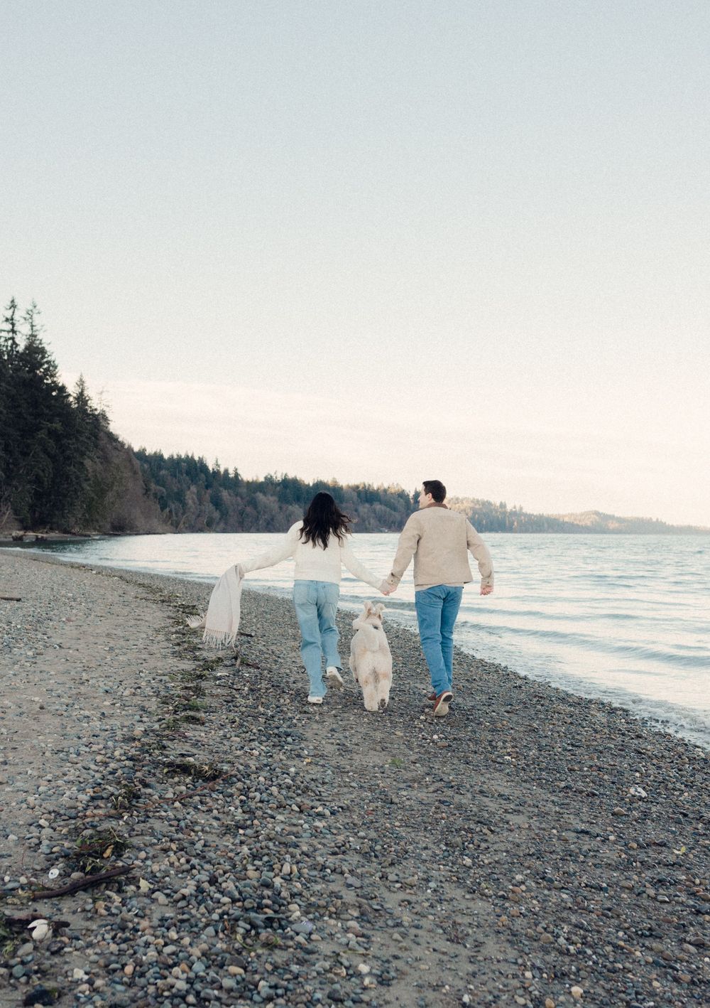 A couple walks along a rocky beach shoreline with evergreen trees in the distance during sunset.