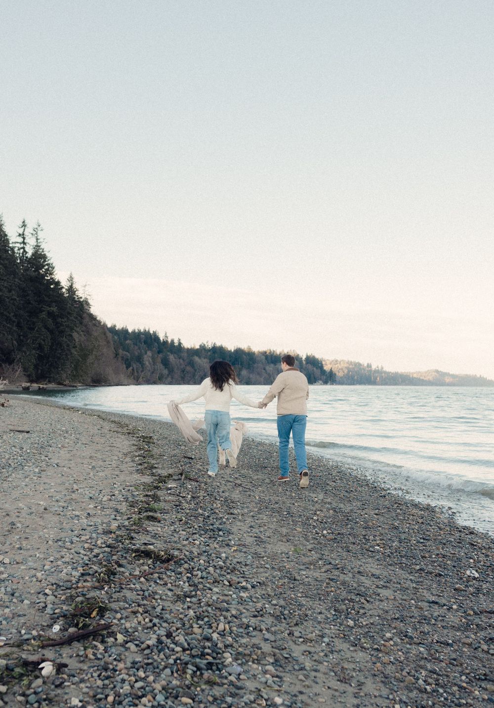 A couple walks along a rocky beach shoreline with evergreen trees in the background during sunset.