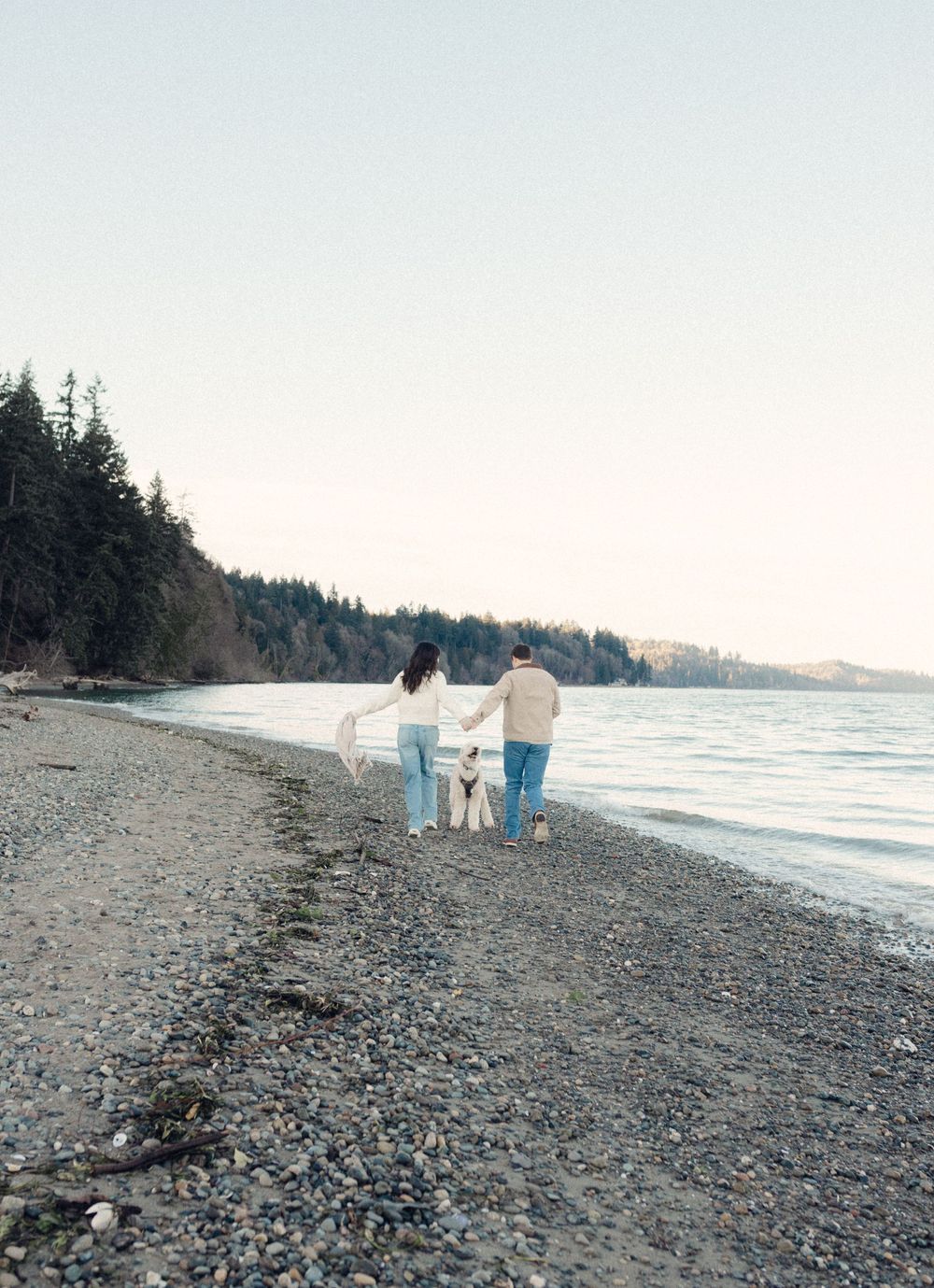 Three figures stroll on a pebbled beach with forested coastline and calm waters in the background.