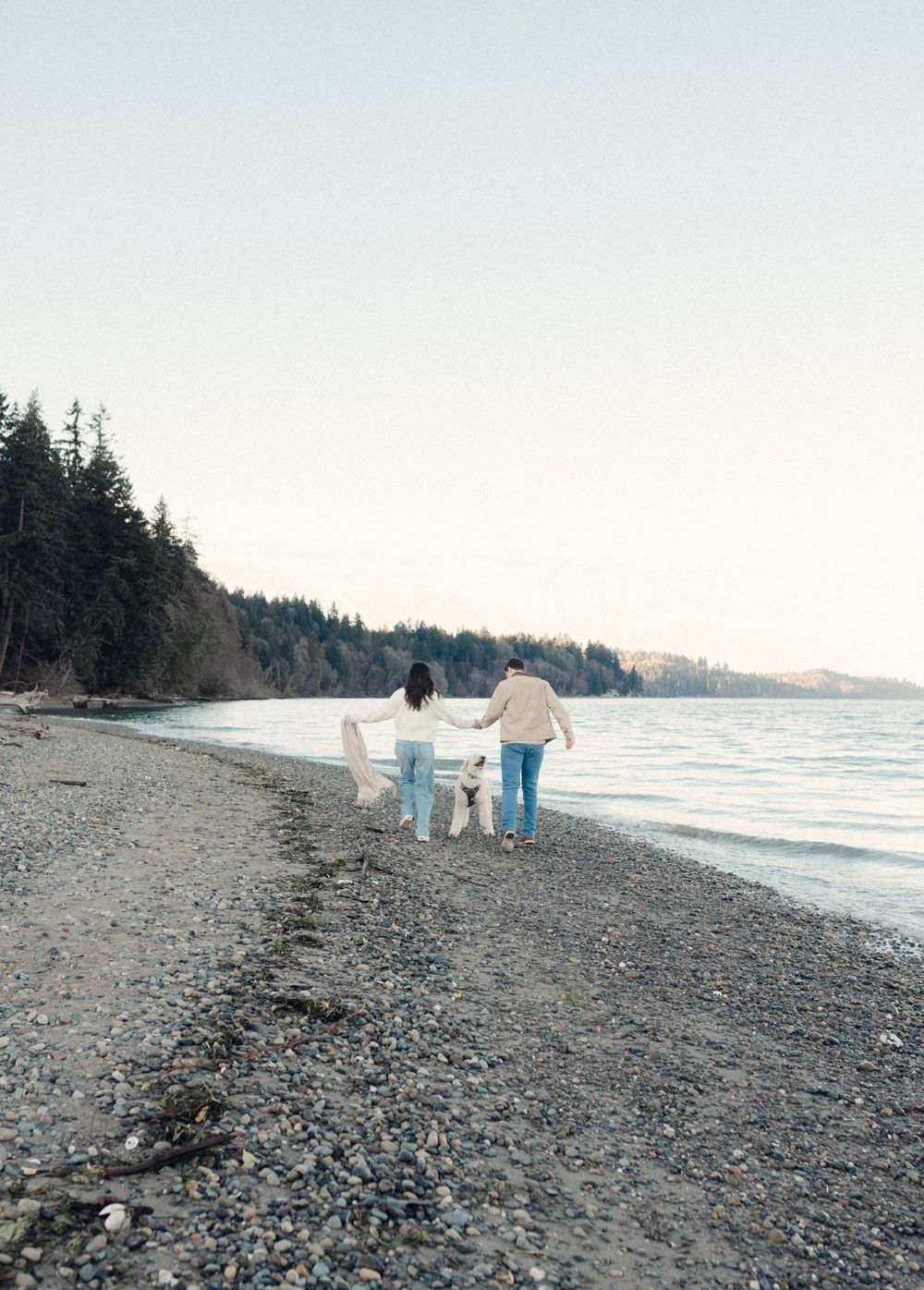 People walking along a rocky beach shoreline with evergreen trees in the background on an overcast day.
